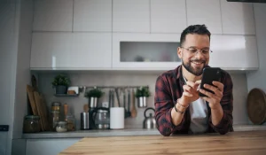 Um homem sorridente está sentado em sua cozinha moderna e usa um smartphone.