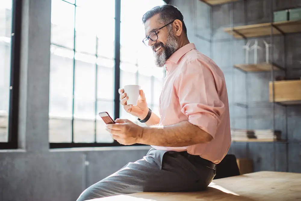 Mature businessman in modern office, using phone and holding a cup of coffee while sitting at a table.