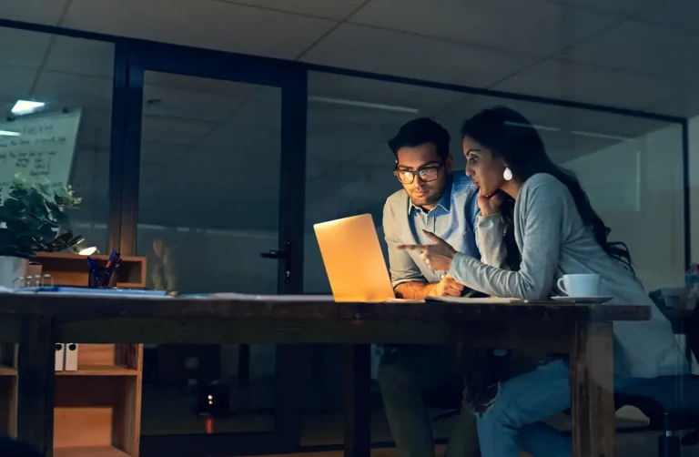 Imagen recortada de dos compañeros trabajando hasta tarde con un portátil en una oficina.