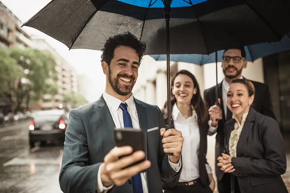 Cheerful businesspeople sharing an umbrella, smiling, and using a smartphone on a rainy city street.