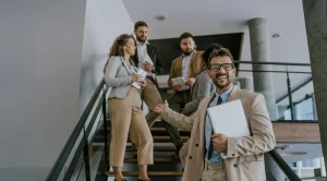 Businessman in tan suit smiling on office staircase, holding laptop, with coworkers in background.