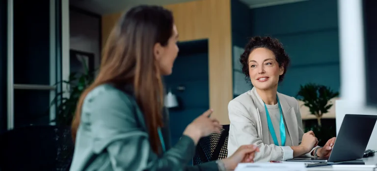 Mature businesswoman working on a computer while talking to female coworker at corporate office.
