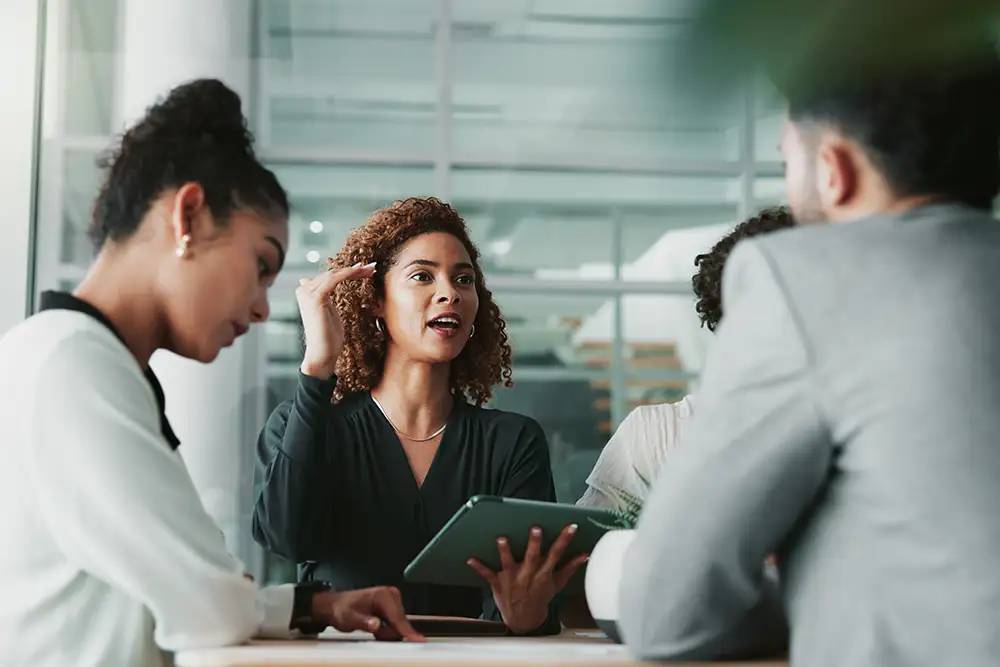Four professionals in a modern office setting engaged in a meeting; a woman in the center is speaking confidently while holding a tablet, with colleagues listening attentively.