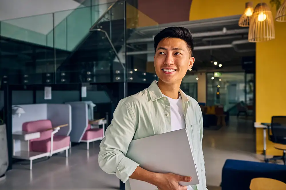 business-goals-2186780917 Portrait of a cheerful young asian man holding a laptop in a modern office environment